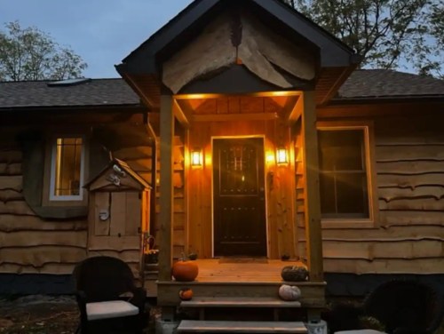 A cozy log cabin entrance illuminated by warm lights, with pumpkins and chairs on the porch.
