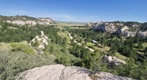 A panoramic view of a lush green valley surrounded by rocky cliffs and a winding road under a clear blue sky.