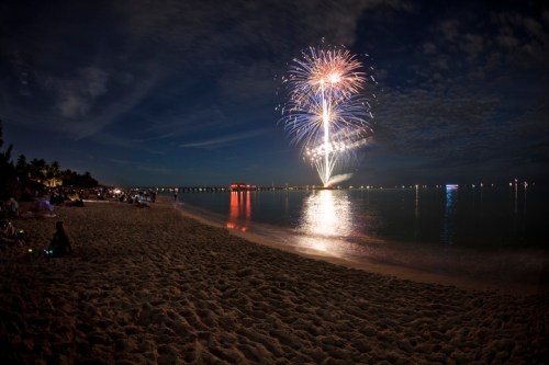 Fireworks illuminate the night sky over a beach, with spectators enjoying the view along the sandy shore.