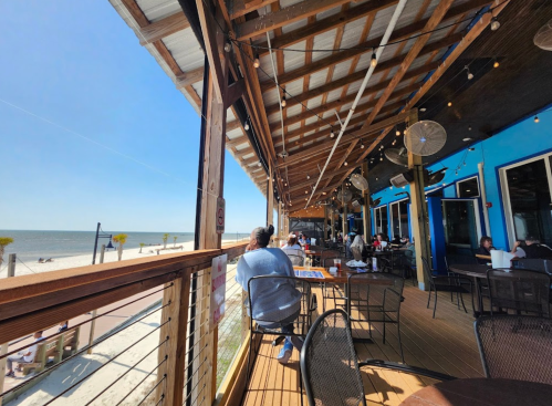 A sunny beachside restaurant patio with diners, overlooking the ocean and a clear blue sky.