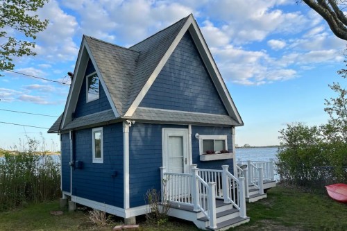 A small blue A-frame house by the water, featuring a porch and surrounded by greenery under a partly cloudy sky.