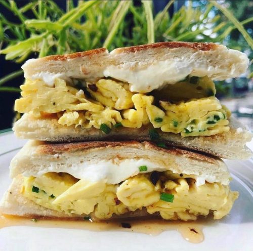 A close-up of a toasted sandwich filled with fluffy scrambled eggs and a dollop of cream, garnished with chives.