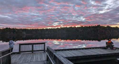 Two people stand on a wooden dock by a calm lake, reflecting colorful clouds at sunset. Trees line the horizon.