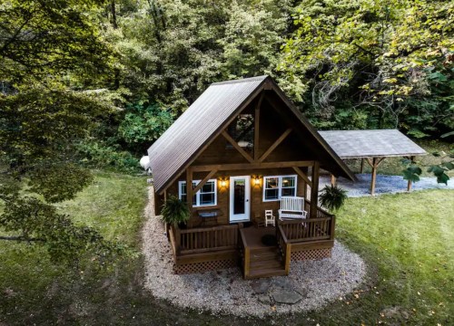 Aerial view of a cozy wooden cabin surrounded by trees, featuring a porch and a gravel path.