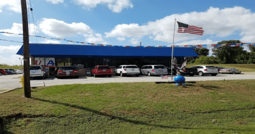 A cafe with a blue roof, several parked cars, and an American flag in front, set against a cloudy sky.