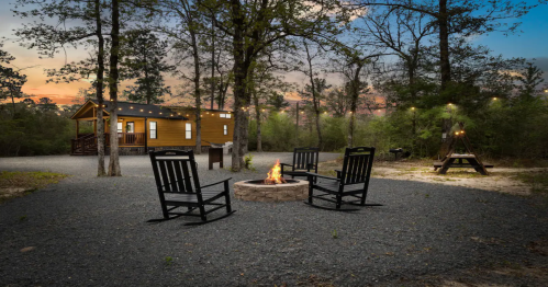 A cozy outdoor setting with four rocking chairs around a fire pit, surrounded by trees and a cabin in the background.