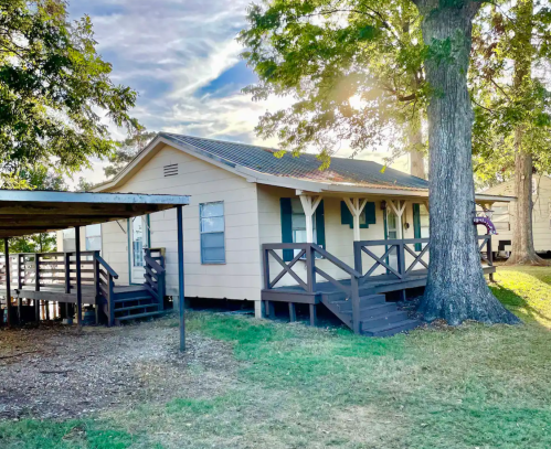 A cozy, light-colored cabin with a porch, surrounded by trees and grass under a bright sky.
