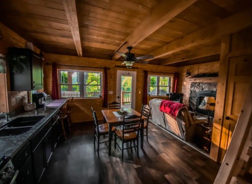 Cozy cabin interior with wooden beams, a dining table, and a living area featuring a stone fireplace and large windows.