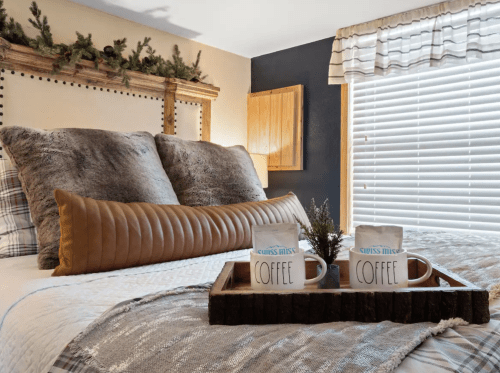 Cozy bedroom with a bed, decorative pillows, and two coffee mugs on a wooden tray. Natural light from a window.
