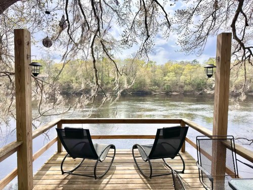 A serene riverside deck with two chairs, surrounded by trees and reflecting water under a cloudy sky.