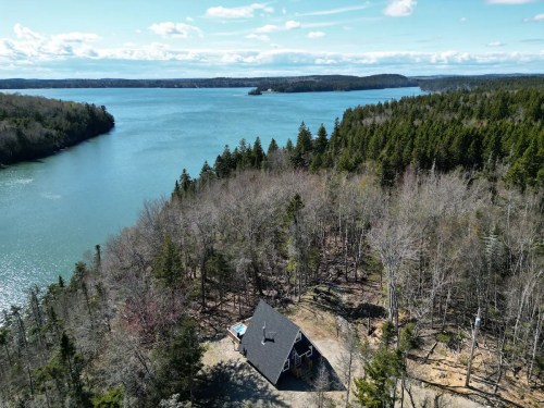 Aerial view of a house by a serene lake, surrounded by trees and hills under a clear blue sky.