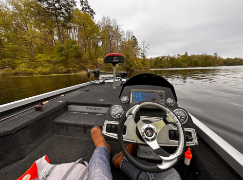 A view from a boat's cockpit, showing a person’s feet and hands on the steering wheel, with calm water and trees in the background.