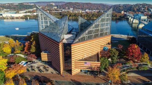 A modern building with a unique triangular roof, surrounded by colorful autumn trees and a river in the background.