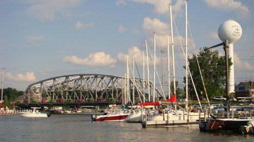A marina with various boats docked, a bridge in the background, and a water tower nearby under a blue sky.