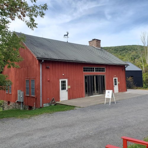 A red barn-style building with large windows, a weather vane, and a gravel path in a scenic outdoor setting.