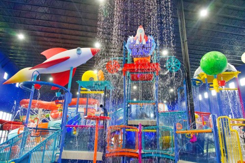 Colorful indoor water park with a large water play structure, featuring a rocket, planets, and cascading water.