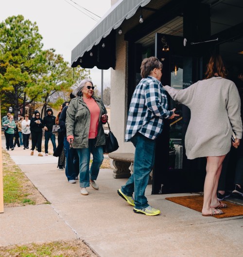 A line of people waiting to enter a store on a cloudy day, with some wearing jackets and casual attire.