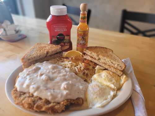 A plate with chicken fried steak topped with gravy, hash browns, two eggs, toast, and condiments of ketchup and hot sauce.