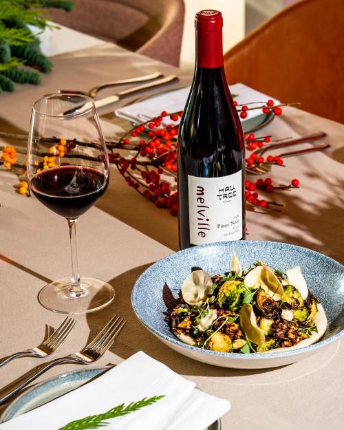A plate of roasted vegetables garnished with herbs, a glass of red wine, and a bottle of Pinot Noir on a dining table.