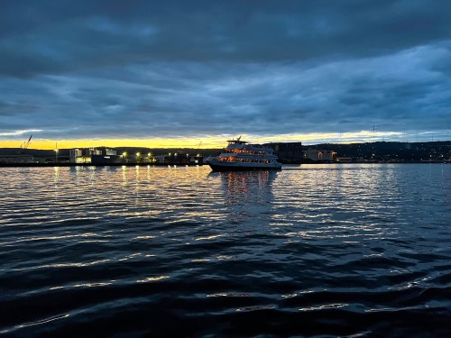 A boat on calm water at sunset, with a cloudy sky and city lights reflecting on the surface.