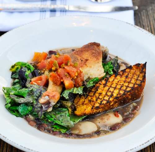 A plate of grilled chicken with cornbread, greens, and a tomato topping served over a bed of black-eyed peas.