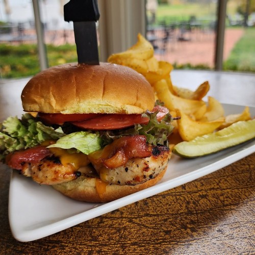 A grilled chicken sandwich with lettuce, tomato, bacon, and cheese, served with fries and a pickle on a plate.