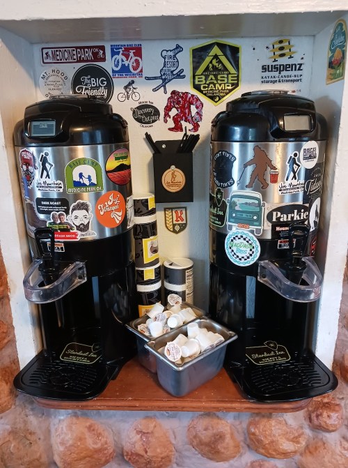 Two coffee dispensers with various stickers on the wall behind them, alongside a container of coffee pods.