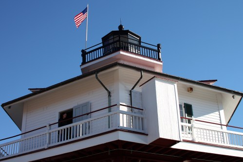 A lighthouse with a flag on top, featuring a balcony and windows against a clear blue sky.