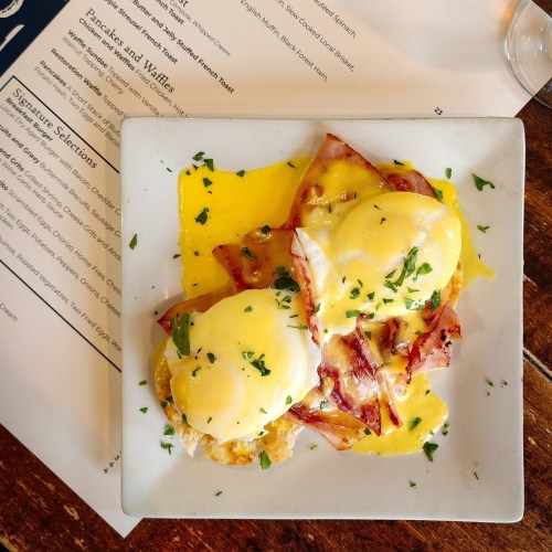 A plate of eggs Benedict with ham, topped with hollandaise sauce and garnished with parsley, next to a menu.