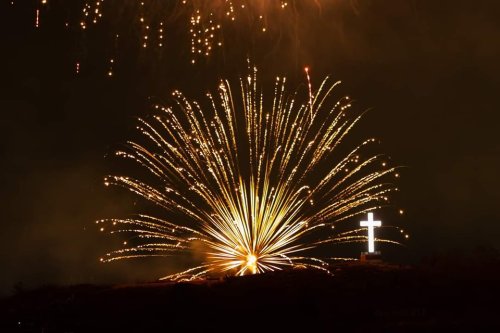 A bright cross illuminated by vibrant fireworks against a dark night sky.