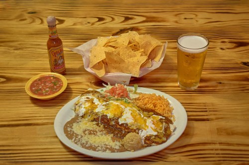 A plate of enchiladas with rice and beans, accompanied by chips, salsa, a bottle of hot sauce, and a glass of beer.