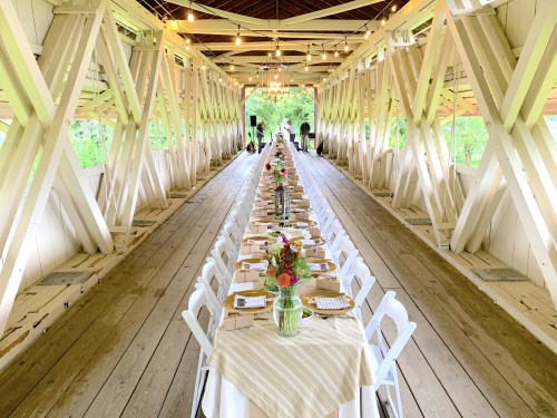 A long dining table set with flowers and tableware inside a rustic, well-lit covered bridge.