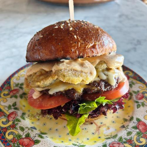 A close-up of a gourmet burger with a sesame seed bun, lettuce, tomato, pickles, and a creamy sauce on a decorative plate.