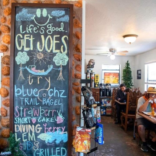 Chalkboard menu in a cozy café, featuring food items and a welcoming message. Customers enjoy their meals in the background.