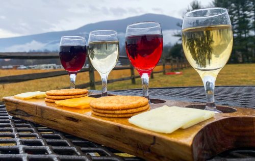 A wooden tray holds four glasses of wine and a selection of cheese and crackers, with mountains in the background.