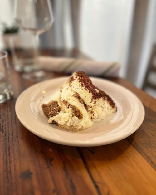 A slice of tiramisu on a white plate, resting on a wooden table, with glasses and a napkin in the background.