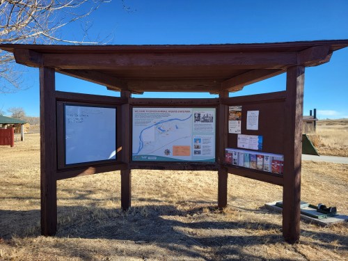 A wooden information kiosk in a park, featuring a map, bulletin board, and brochures, set against a clear blue sky.