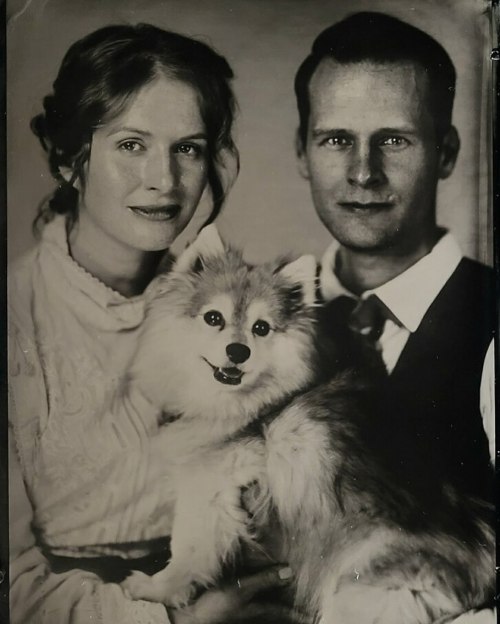 A vintage portrait of a couple holding a fluffy dog, both smiling warmly at the camera.