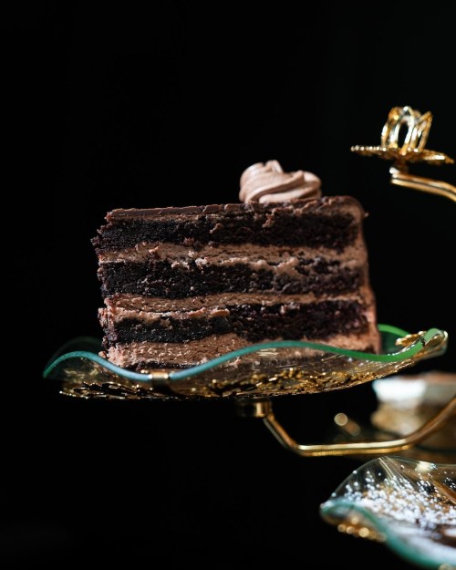 A slice of rich chocolate cake with layers of frosting, displayed on an ornate glass stand against a dark background.