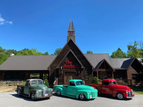 Three vintage trucks in front of a rustic building with a clock tower, labeled "The Abbey," under a clear blue sky.