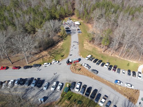 Aerial view of a gathering in a wooded area, with cars parked along the road and a group of people near a shelter.