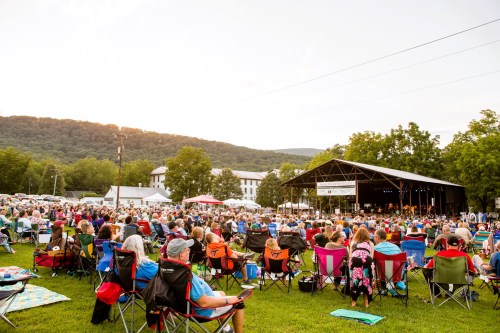 A large crowd sits in colorful chairs at an outdoor concert, with a stage and mountains in the background.