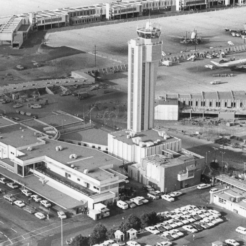 Aerial view of an airport terminal and control tower, with parked planes and vehicles in black and white.