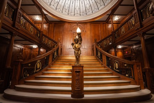 Elegant wooden staircase with a statue holding a lamp, illuminated by a large dome ceiling. Richly detailed decor.