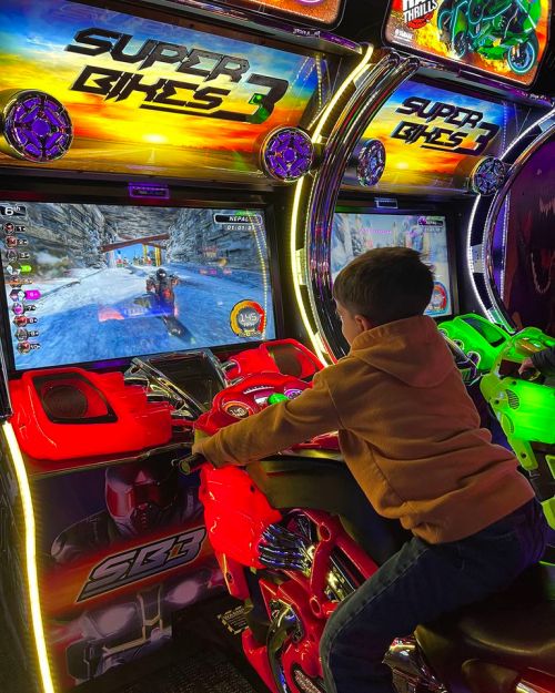 A child plays a motorcycle racing arcade game, focused on the screen with vibrant graphics and colorful game machines.