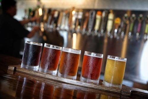 A flight of five beer samples in clear glasses on a wooden tray, with a bar and taps in the background.