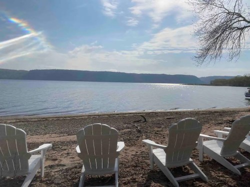 A serene lakeside view with white Adirondack chairs facing calm water and distant hills under a partly cloudy sky.