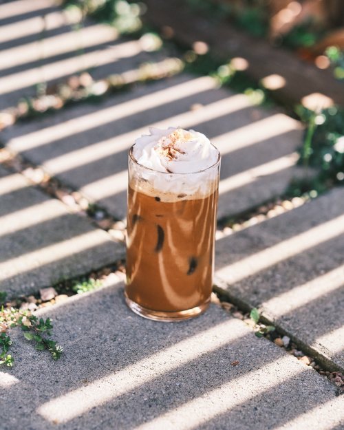 A glass of iced coffee topped with whipped cream, sitting on a stone surface with shadows from nearby slats.