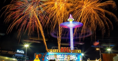 Colorful fireworks illuminate a carnival scene featuring a prize booth and a tall, lit tower at night.