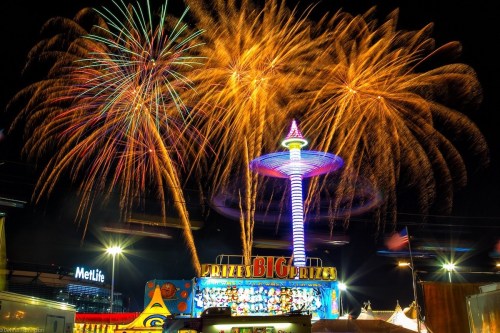 Colorful fireworks illuminate the night sky above a carnival with a tall, lit tower and a prize booth.